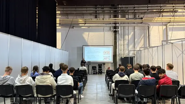 Eine Gruppe von Schülern sitzt in einem großen Raum auf Stühlen, während eine Präsentation auf einer Leinwand gezeigt wird. Im Vordergrund steht ein Referent, der Informationen vermittelt. Der Raum ist einfach und industriell gestaltet.