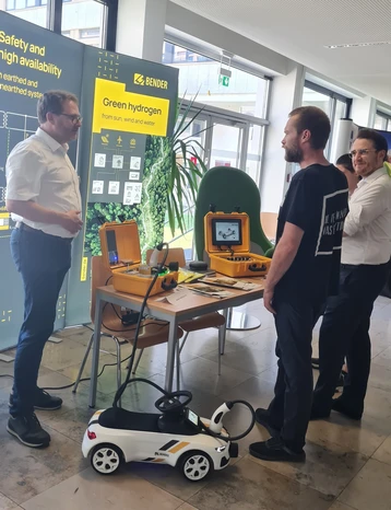 Drei Personen stehen an einem Messestand. Auf dem Tisch sind Geräte in gelben Koffer und ein Display sichtbar. Im Hintergrund ist ein Banner mit Informationen zu grünem Wasserstoff zu sehen. Die Personen scheinen eine Präsentation oder Diskussion zu führen.