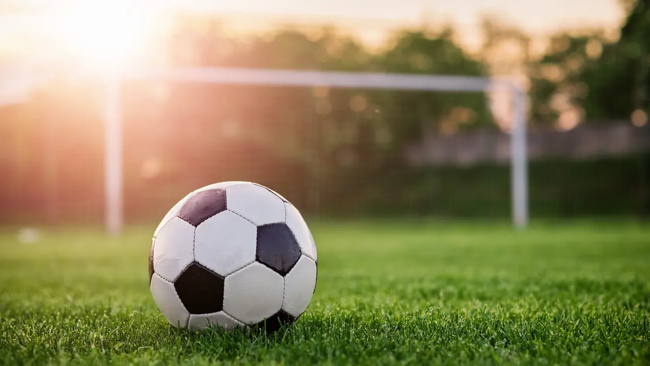 Ein Fußball liegt auf dem grünen Rasen vor einem Tor. Die Sonne scheint im Hintergrund und sorgt für eine warme Atmosphäre. Das Bild vermittelt ein Gefühl von Sport und Freizeit auf einem Fußballplatz.