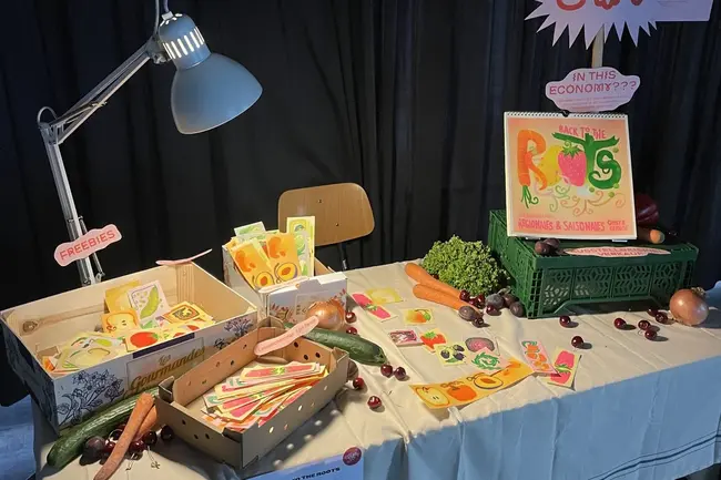 Ein Tisch mit mehreren Kisten, die mit bunten, handgezeichneten Lebensmittelkarten gefüllt sind, darunter Gemüse und Obst. Eine Lampe beleuchtet den Tisch, auf dem auch frische Karotten, Trauben und eine Schüssel mit Gemüse liegen. Ein Schild fordert zur Teilnahme auf.