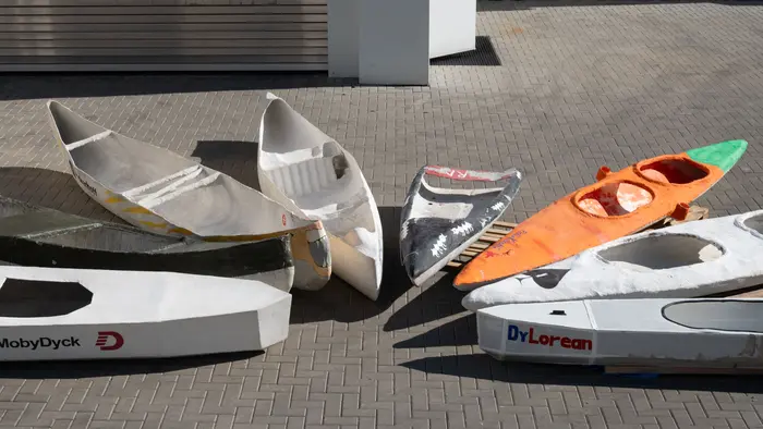 Mehrere leere Bootshüllen liegen auf einem Pflasterboden. Die Boote sind in verschiedenen Farben und Designs, einige mit Aufschriften wie „MobyDyck“ und „DrLorean“. Die Anordnung zeigt eine Vielzahl von Formen und Größen. Das Bild vermittelt eine kreative, handwerkliche Atmosphäre.