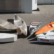 Mehrere leere Bootshüllen liegen auf einem Pflasterboden. Die Boote sind in verschiedenen Farben und Designs, einige mit Aufschriften wie „MobyDyck“ und „DrLorean“. Die Anordnung zeigt eine Vielzahl von Formen und Größen. Das Bild vermittelt eine kreative, handwerkliche Atmosphäre.