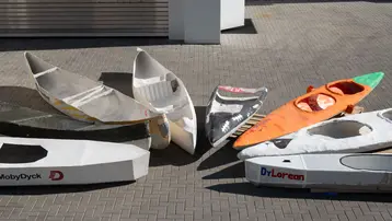 Mehrere leere Bootshüllen liegen auf einem Pflasterboden. Die Boote sind in verschiedenen Farben und Designs, einige mit Aufschriften wie „MobyDyck“ und „DrLorean“. Die Anordnung zeigt eine Vielzahl von Formen und Größen. Das Bild vermittelt eine kreative, handwerkliche Atmosphäre.