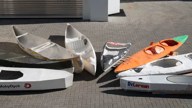 Mehrere leere Bootshüllen liegen auf einem Pflasterboden. Die Boote sind in verschiedenen Farben und Designs, einige mit Aufschriften wie „MobyDyck“ und „DrLorean“. Die Anordnung zeigt eine Vielzahl von Formen und Größen. Das Bild vermittelt eine kreative, handwerkliche Atmosphäre.