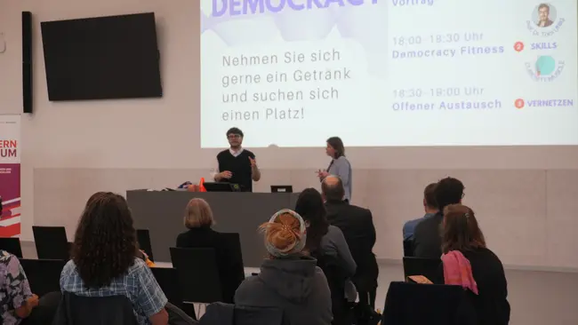 Eine Präsentation im Rahmen einer Veranstaltung mit dem Titel "Doing Democracy". Zwei Personen stehen an einem Tisch, während im Hintergrund Informationen zu Vorträgen und Aktivitäten eingeblendet werden. Der Raum ist mit mehreren Teilnehmern besetzt, die auf die Präsentation achten.