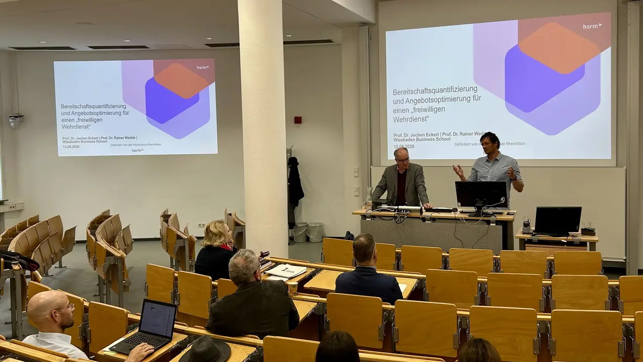 In einem Seminarraum präsentieren zwei Referenten vor einem Publikum. Auf einem Bildschirm ist eine Folie mit dem Titel „Betreuungsgestaltung und Angebotsgestaltung für einen zweiten Wiederstart“ zu sehen. Die Teilnehmer sitzen in Reihen, einige haben Notebooks oder Notizen vor sich.
