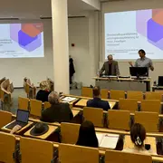 In einem Seminarraum präsentieren zwei Referenten vor einem Publikum. Auf einem Bildschirm ist eine Folie mit dem Titel „Betreuungsgestaltung und Angebotsgestaltung für einen zweiten Wiederstart“ zu sehen. Die Teilnehmer sitzen in Reihen, einige haben Notebooks oder Notizen vor sich.