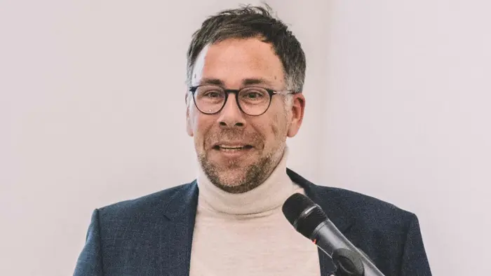 Ein Mann mit Brille und kurzem Haar trägt einen hellen Pullover unter einem dunklen Blazer. Er steht an einem Rednerpult und hält ein Mikrofon. Der Mann spricht, während er mit der Hand gestikuliert. Hintergrund ist hell und schlicht.