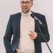 Ein Mann mit Brille und kurzem Haar trägt einen hellen Pullover unter einem dunklen Blazer. Er steht an einem Rednerpult und hält ein Mikrofon. Der Mann spricht, während er mit der Hand gestikuliert. Hintergrund ist hell und schlicht.