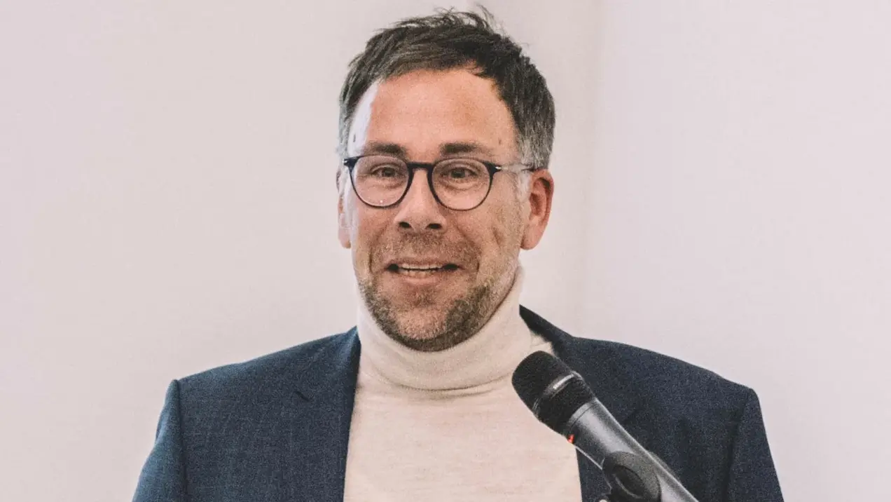 Ein Mann mit Brille und kurzem Haar trägt einen hellen Pullover unter einem dunklen Blazer. Er steht an einem Rednerpult und hält ein Mikrofon. Der Mann spricht, während er mit der Hand gestikuliert. Hintergrund ist hell und schlicht.