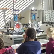 In einem Laborraum sitzen mehrere Schüler mit dem Rücken zur Kamera. Vor ihnen befinden sich Apparaturen zur Durchführung von Experimenten. Lichtstrahlen fallen durch Fenster und beleuchten die Szene. Der Raum ist mit verschiedenen technischen Geräten ausgestattet.