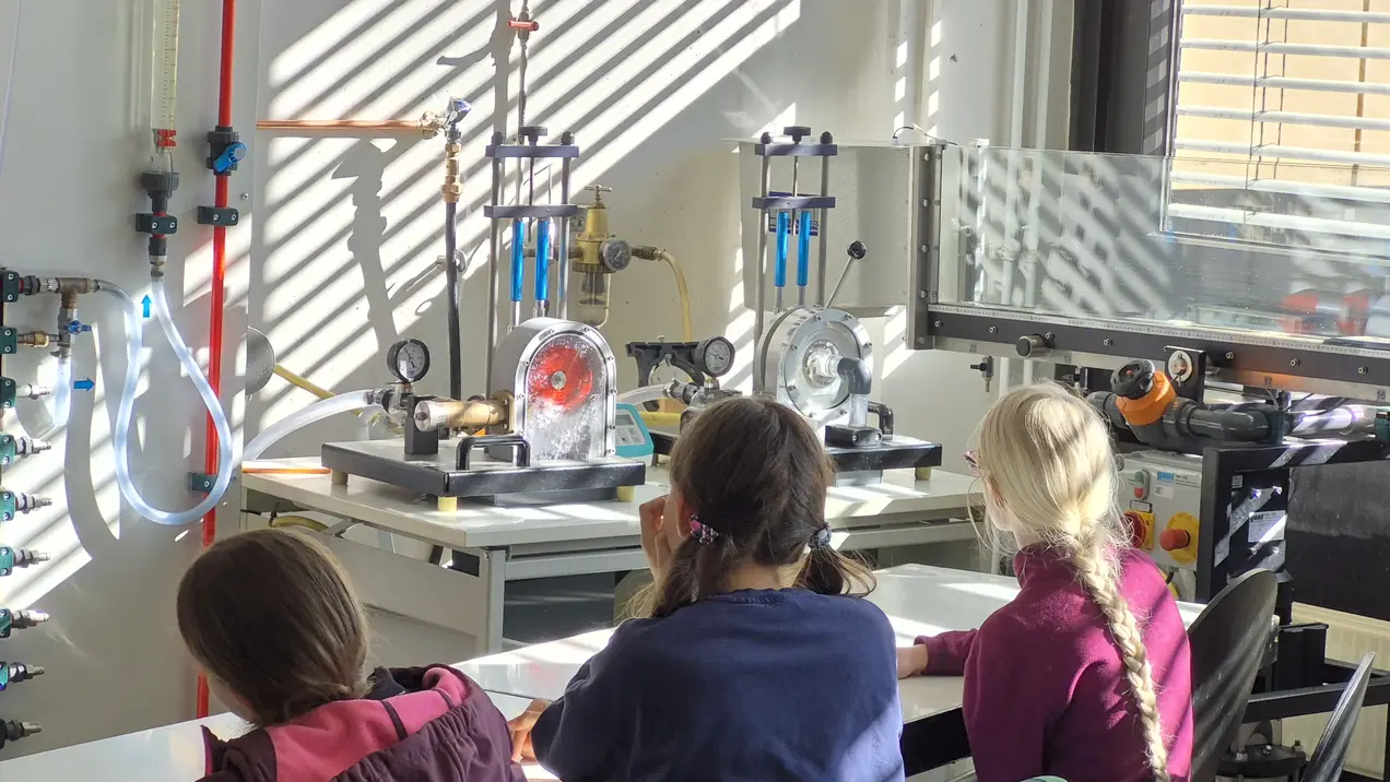 In einem Laborraum sitzen mehrere Schüler mit dem Rücken zur Kamera. Vor ihnen befinden sich Apparaturen zur Durchführung von Experimenten. Lichtstrahlen fallen durch Fenster und beleuchten die Szene. Der Raum ist mit verschiedenen technischen Geräten ausgestattet.