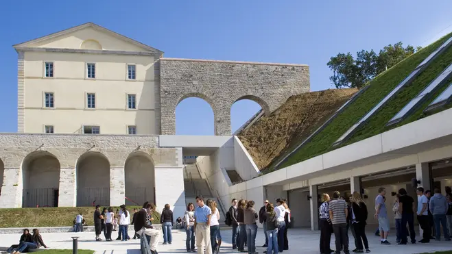 Eine Gruppe von Menschen versammelt sich im Freien vor einem historischen Gebäude mit einem großen, offenen Bogen. Im Hintergrund sind moderne architektonische Elemente und eine grüne, bewachsene Fläche. Der Himmel ist klar und blau.