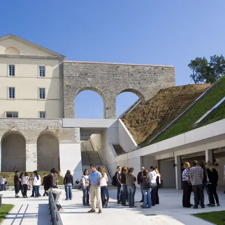 Eine Gruppe von Menschen versammelt sich im Freien vor einem historischen Gebäude mit einem großen, offenen Bogen. Im Hintergrund sind moderne architektonische Elemente und eine grüne, bewachsene Fläche. Der Himmel ist klar und blau.