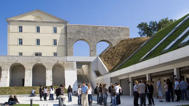 Eine Gruppe von Menschen versammelt sich im Freien vor einem historischen Gebäude mit einem großen, offenen Bogen. Im Hintergrund sind moderne architektonische Elemente und eine grüne, bewachsene Fläche. Der Himmel ist klar und blau.