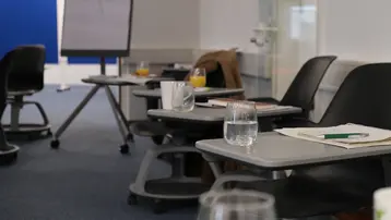 Ein Besprechungsraum mit mehreren Tischen und Stühlen ist abgebildet. Auf den Tischen stehen Gläser mit Wasser, eine Tasse und einige Unterlagen. Im Hintergrund ist ein Flipchart zu sehen, das auf einen Workshop oder eine Schulung hinweist.