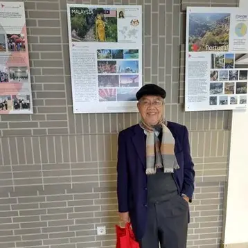 Ein älterer Mann in einem dunkelblauen Mantel und einer gestreiften Schal steht lächelnd und hält eine rote Einkaufstasche. Im Hintergrund sind Informationsplakate über Malaysia und Portugal an der Wand angebracht. Der Boden ist hell und die Umgebung ist gut beleuchtet.