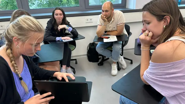 Sechs junge Menschen sitzen in einem Raum und diskutieren. Eine Person auf der linken Seite arbeitet an einem Laptop, während die anderen aufmerksam zuhören und nachdenklich wirken. Der Raum hat große Fenster, die Tageslicht hereinlassen.