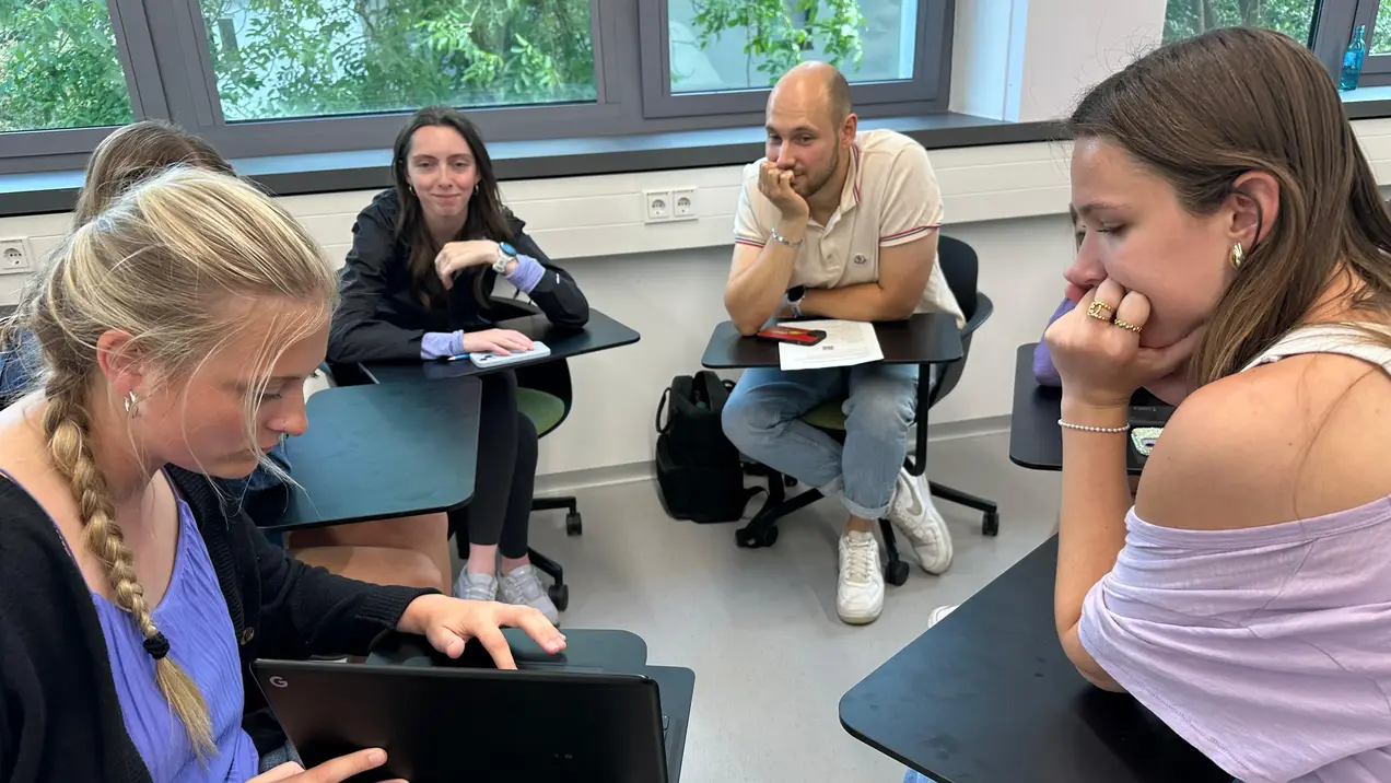 Sechs junge Menschen sitzen in einem Raum und diskutieren. Eine Person auf der linken Seite arbeitet an einem Laptop, während die anderen aufmerksam zuhören und nachdenklich wirken. Der Raum hat große Fenster, die Tageslicht hereinlassen.