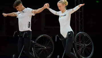 Zwei Sportler stehen auf einer speziellen Fahrradbühne, während sie sich an den Händen halten und ihre Arme ausbreiten. Sie führen eine anspruchsvolle akrobatische Darbietung vor, die Gleichgewicht und Teamarbeit erfordert. Beide tragen sportliche Kleidung in unterschiedlichen Designs.
