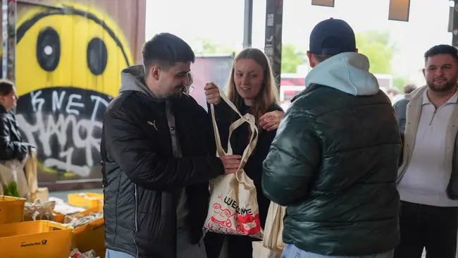 Drei Personen stehen in einem Raum mit Graffiti an der Wand. Zwei Männer und eine Frau interagieren, wobei die Frau eine Tragetasche zeigt. Im Hintergrund sind orangefarbene Kisten sichtbar, die auf eine geschäftige Umgebung hindeuten.