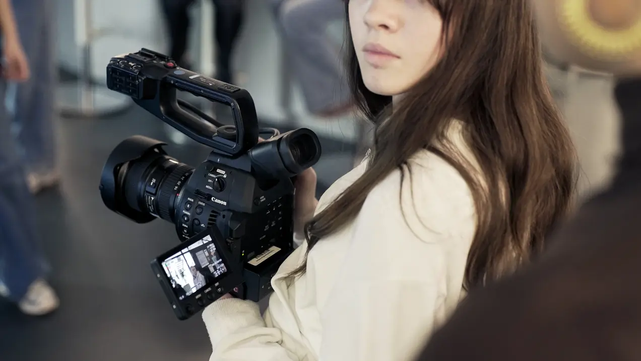 Eine Person mit langen, braunen Haaren hält eine professionelle Kamera in der Hand. Die Kamera hat ein großes Objektiv und ein Display, auf dem eine Aufnahme zu sehen ist. Die Umgebung deutet auf eine Veranstaltung oder einen kreativen Kontext hin.