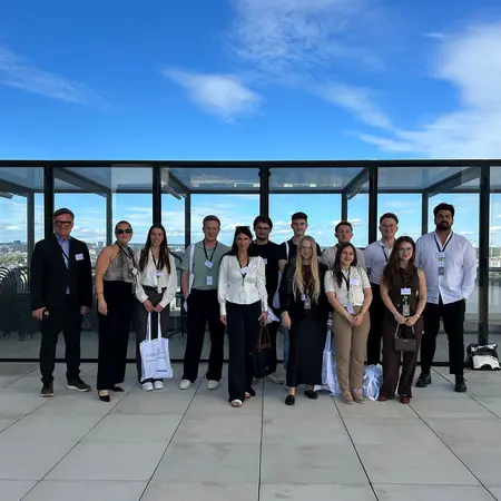 Eine Gruppe von 14 Personen steht auf einer Terrasse mit Glaswänden. Sie tragen formelle Kleidung und lächeln in die Kamera. Im Hintergrund ist ein blauer Himmel zu sehen. Die Menschen scheinen an einer Veranstaltung oder einem Treffen teilzunehmen.