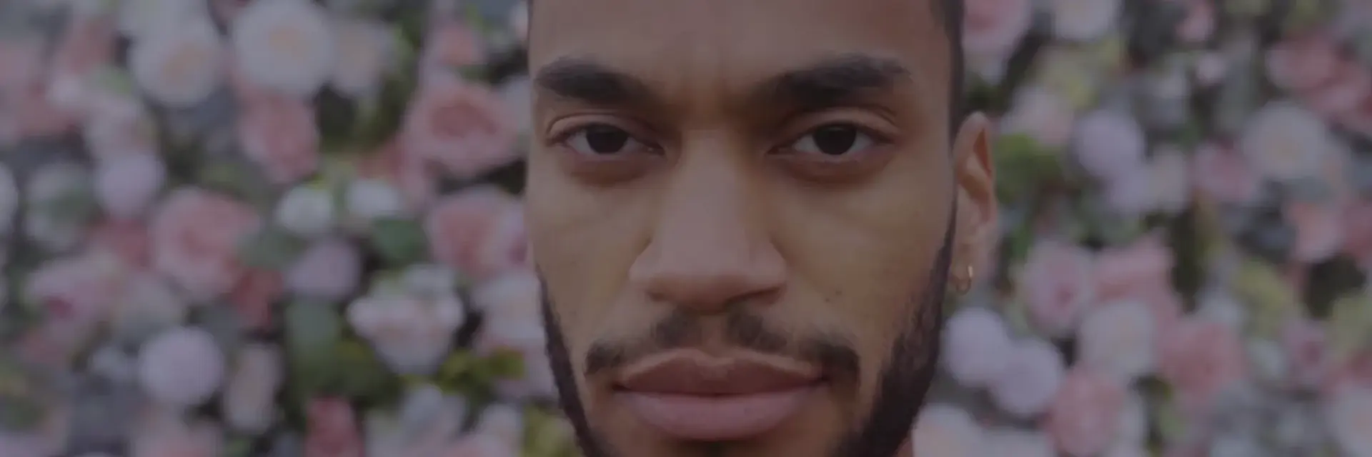 Ein junger Mann mit Bart und Kurzhaarschnitt blickt direkt in die Kamera. Er trägt ein dunkelrotes Shirt. Im Hintergrund ist eine Wand aus bunten, blühenden Blumen zu sehen, die eine romantische Atmosphäre schafft.