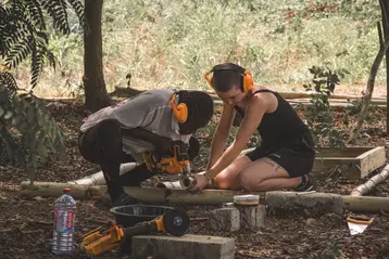 Two students are working together outdoors on a bamboo project. One person is using a machine while the other assists. Both are wearing ear protection and are surrounded by green countryside. Tools and materials are scattered on the ground.