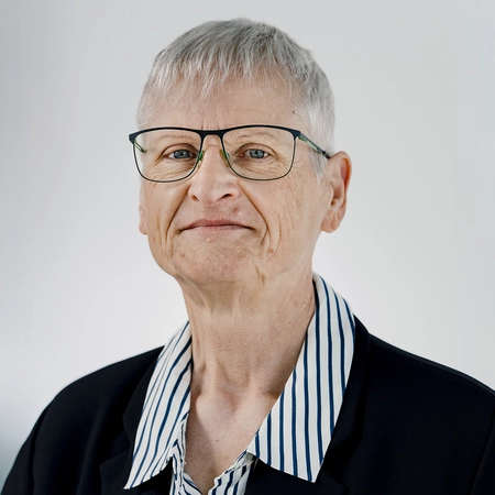 Ein älterer, lächelnder Mensch mit kurzen, grauen Haaren und Brille trägt ein schwarzes Sakko über einem gestreiften Hemd. Der Hintergrund ist schlicht und hell.