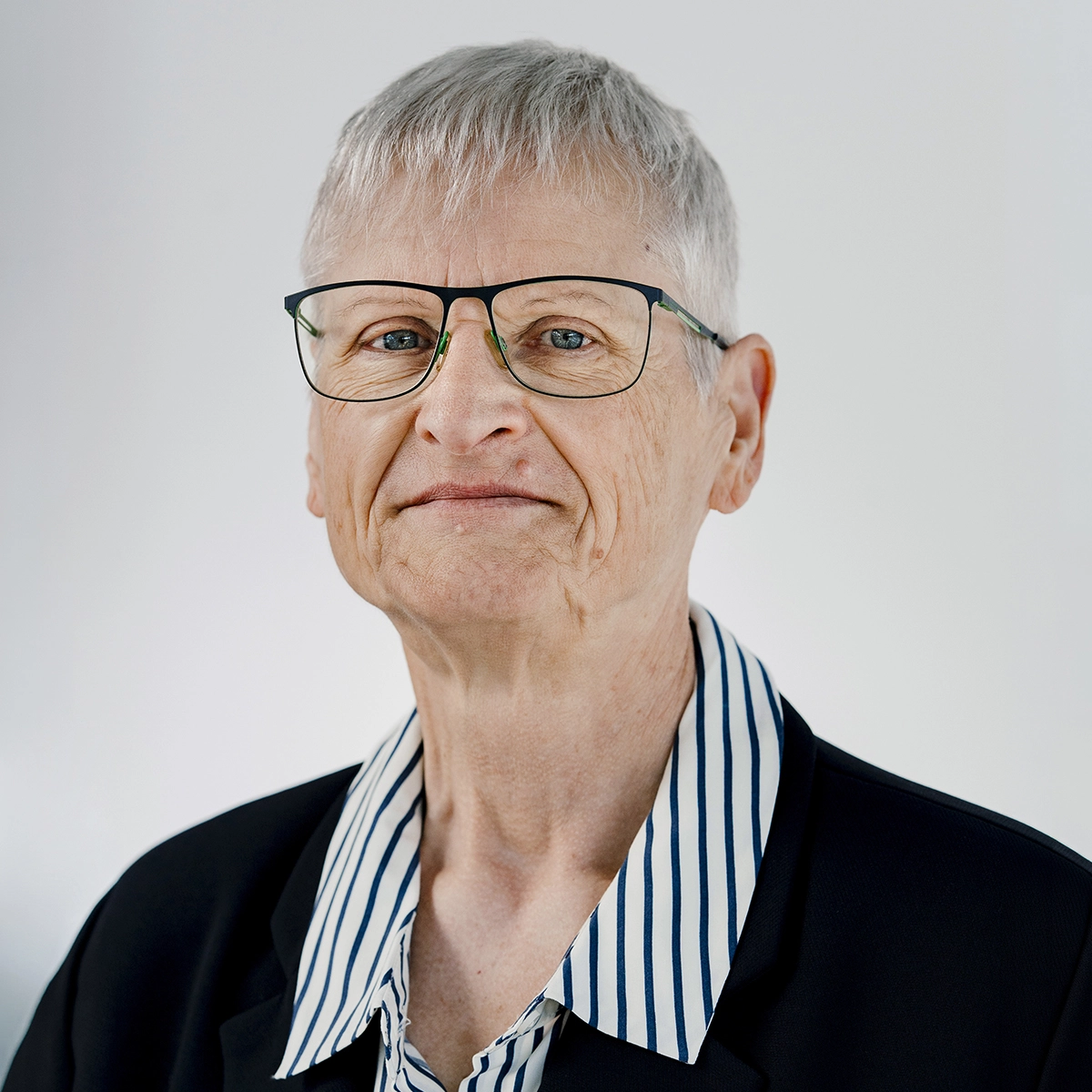 Ein älterer, lächelnder Mensch mit kurzen, grauen Haaren und Brille trägt ein schwarzes Sakko über einem gestreiften Hemd. Der Hintergrund ist schlicht und hell.