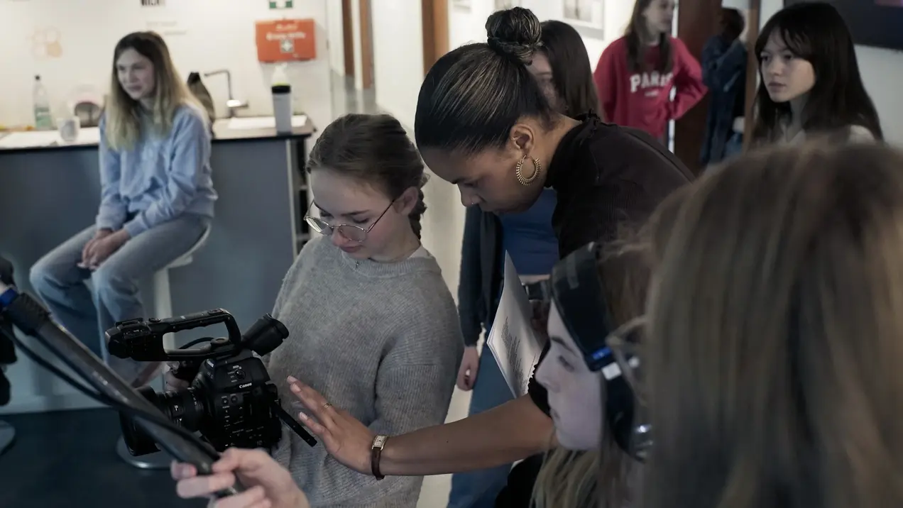 Eine Gruppe von jungen Frauen steht zusammen in einem Raum. Eine Person zeigt einer anderen, wie man eine Kamera bedient. Die Atmosphäre ist konzentriert und neugierig, während die Teilnehmerinnen lernen und sich austauschen.