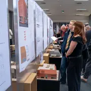 In einer Ausstellung betrachten Besucher Architekturzeichnungen und Modelle. Eine Frau schaut sich Pläne an. Weitere Personen sind im Hintergrund mit verschiedenen Zeichnungen und Modellen beschäftigt, was das Interesse an zeitgenössischer Architektur zeigt. Die Modelle und Pläne sind Abschlussarbeiten im Studiengang Architektur an der Hochschule RheinMain.