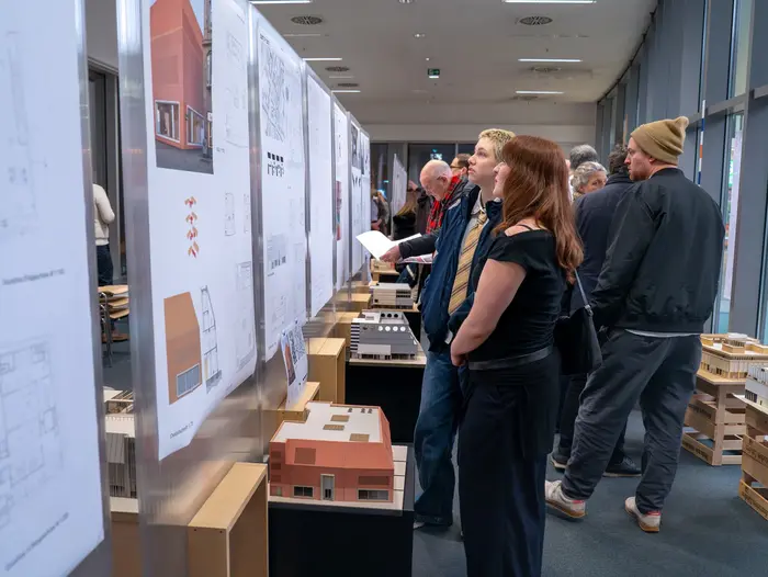 In einer Ausstellung betrachten Besucher Architekturzeichnungen und Modelle. Eine Frau schaut sich Pläne an. Weitere Personen sind im Hintergrund mit verschiedenen Zeichnungen und Modellen beschäftigt, was das Interesse an zeitgenössischer Architektur zeigt. Die Modelle und Pläne sind Abschlussarbeiten im Studiengang Architektur an der Hochschule RheinMain.