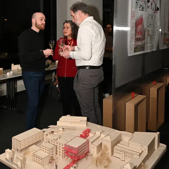 Eine Gruppe von drei Personen steht im Stadtlabor der Hochschule RheinMain und unterhält sich. Vor ihnen befindet sich ein Architekturmodell, das Gebäude und eine rote Struktur im Mittelpunkt zeigt. Es handelt sich um den Teil einer Thesis im Studiengang Architektur. Im Hintergrund sind Präsentationsmaterialien zu sehen.
