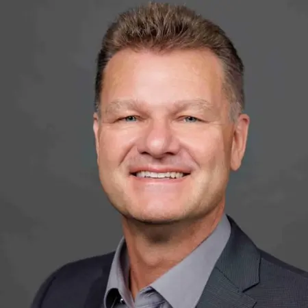 Porträt eines lächelnden Mannes mit kurzem, blondem Haar. Er trägt ein graues Hemd und einen dunklen Anzug. Der Hintergrund ist neutral und grau. Der Mann wirkt freundlich und professionell.