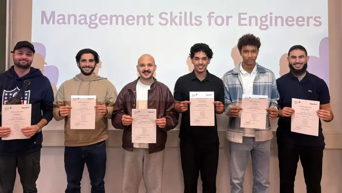 Sechs Teilnehmer halten ihre Zertifikate in die Kamera. Im Hintergrund zeigt ein Bildschirm den Titel „WINTERSCHOOL 2026“ und „Management Skills for Engineers“. Die Atmosphäre ist festlich, und die Teilnehmer scheinen stolz auf ihre Leistungen zu sein.