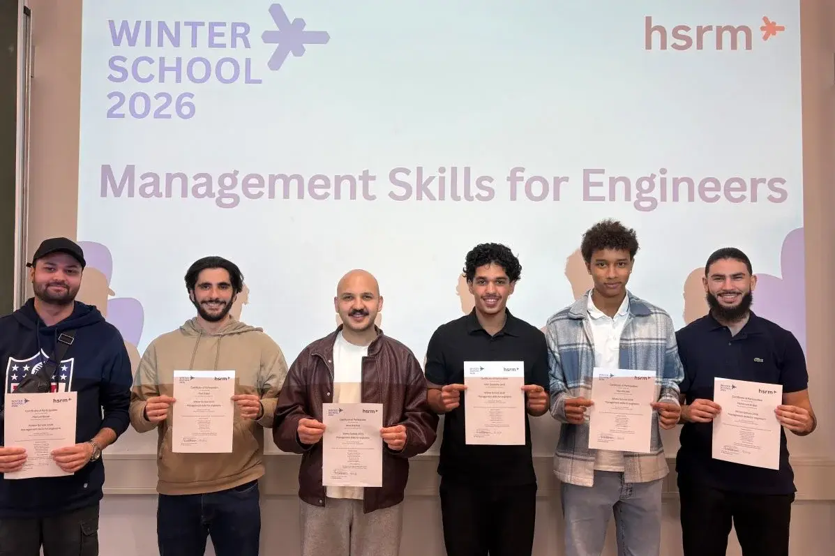 Sechs Teilnehmer halten ihre Zertifikate in die Kamera. Im Hintergrund zeigt ein Bildschirm den Titel „WINTERSCHOOL 2026“ und „Management Skills for Engineers“. Die Atmosphäre ist festlich, und die Teilnehmer scheinen stolz auf ihre Leistungen zu sein.
