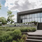 Man sieht ein modernes Gebäude mit Glassfront und ein Schriftzug, der "Hochschule RheinMain" sagt mit dem HSRM Logo (ein weißer Stern) daneben. Der Himmel ist bewölkt und der Rasen vor dem Gebäude grün. Es führen Treppen rechts auf dem Bild zum Kurtschumacher-Ring Campus (KSR).