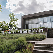 Man sieht ein modernes Gebäude mit Glassfront und ein Schriftzug, der "Hochschule RheinMain" sagt mit dem HSRM Logo (ein weißer Stern) daneben. Der Himmel ist bewölkt und der Rasen vor dem Gebäude grün. Es führen Treppen rechts auf dem Bild zum Kurtschumacher-Ring Campus (KSR).