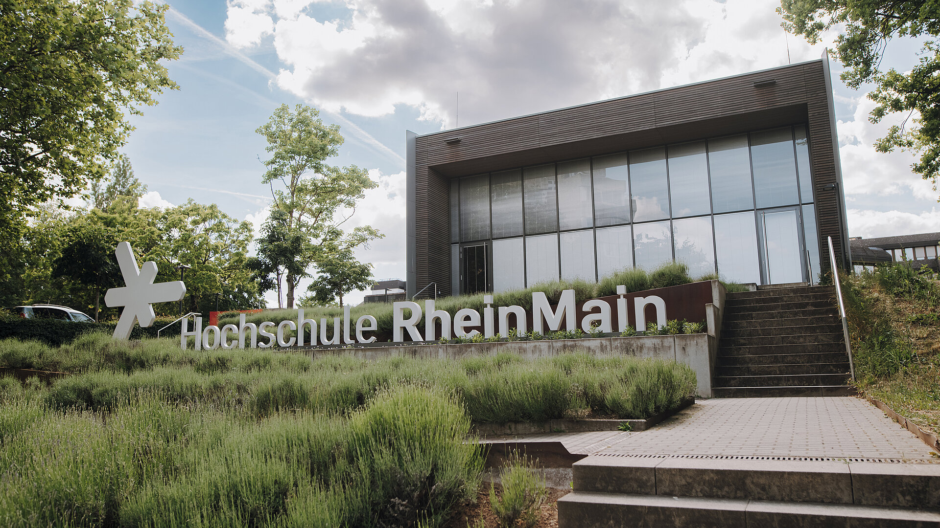 Man sieht ein modernes Gebäude mit Glassfront und ein Schriftzug, der "Hochschule RheinMain" sagt mit dem HSRM Logo (ein weißer Stern) daneben. Der Himmel ist bewölkt und der Rasen vor dem Gebäude grün. Es führen Treppen rechts auf dem Bild zum Kurtschumacher-Ring Campus (KSR).