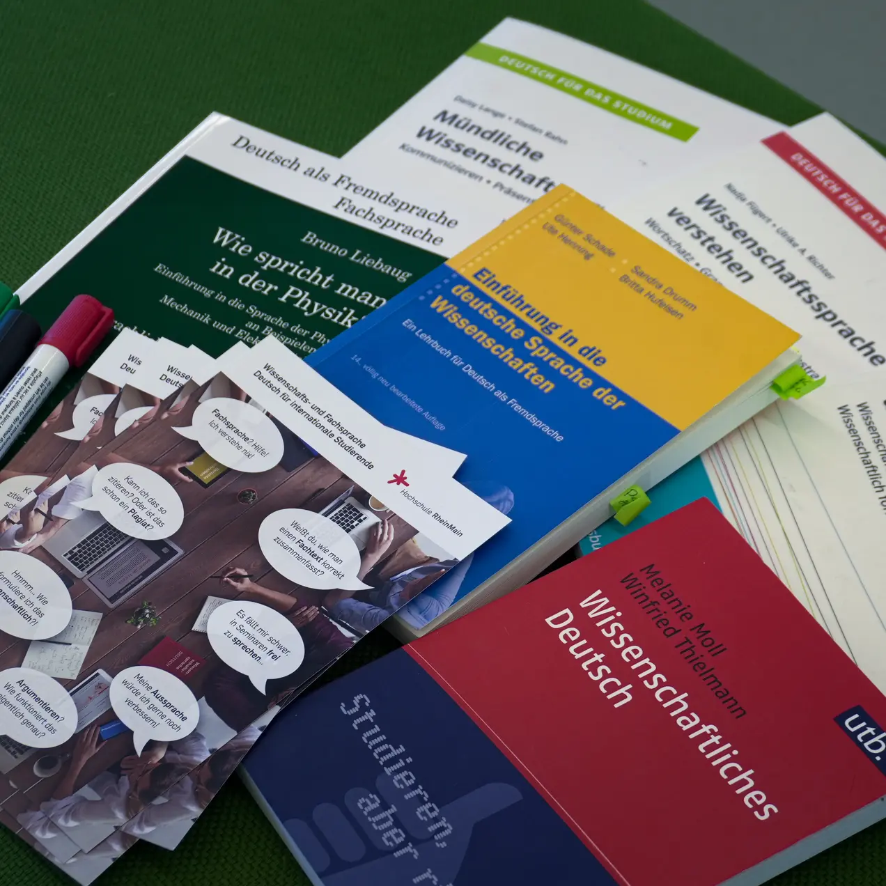 Auf einem grünen Hintergrund liegen verschiedene Bücher und Materialien, darunter Titel über wissenschaftliches Deutsch, wissenschaftliche Sprache und Physik. Außerdem sind mehrere Marker in verschiedenen Farben sichtbar, die auf eine Lern- oder Studierumgebung hinweisen.