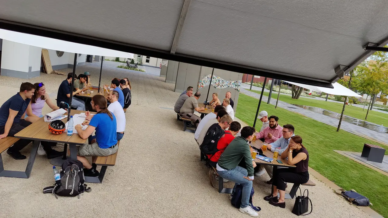 Eine Gruppe von Menschen sitzt an mehreren Tischen im Freien und unterhält sich. Einige Personen essen und arbeiten zusammen, während andere miteinander plaudern. Die Umgebung ist grün und gepflegt, mit Sonnenschirmen.