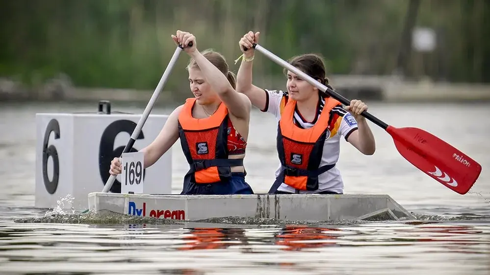 Zwei Mädchen paddeln in einem engen Boot auf einem Gewässer. Beide tragen Schwimmwesten und konzentrieren sich auf das Rudern. Im Hintergrund sind Wettkampfszenen zu erkennen, während die Mädchen gemeinsam versuchen, schnell voranzukommen.