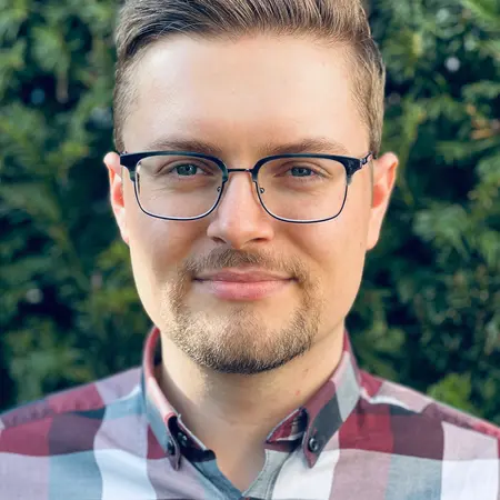Ein junger Mann mit Brille steht vor einem grünen Hintergrund. Er hat kurzes, hellbraunes Haar und trägt ein kariertes Hemd in roten und weißen Farben. Sein Gesichtsausdruck ist freundlich und selbstbewusst.