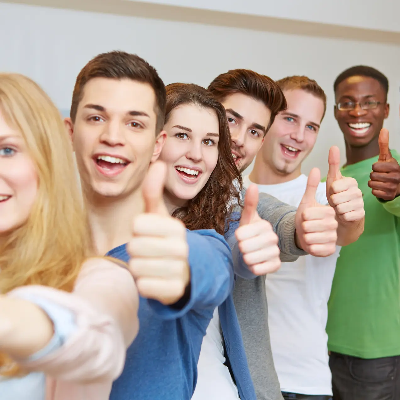 Eine Gruppe von sieben jungen Menschen steht in einer Reihe und lächelt. Alle halten ihre Daumen hoch, was Zustimmung oder Freude ausdrückt. Der Hintergrund ist hell und neutral gestaltet.