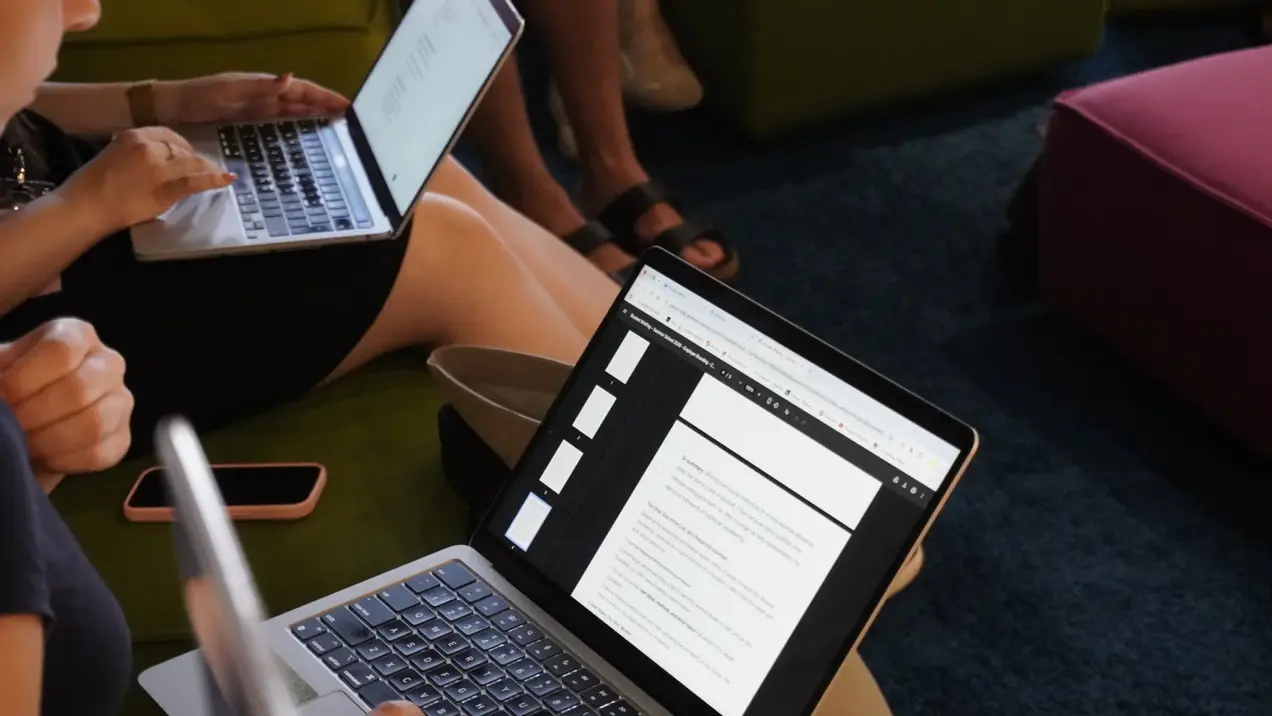 Zwei Personen sitzen auf einer Couch und arbeiten an ihren Laptops. Auf einem Laptop ist ein Dokument mit Text zu sehen. Eine Person hält ein Smartphone in der Hand. Der Hintergrund ist unscharf und zeigt andere Personen, die ebenfalls mit Laptops beschäftigt sind.