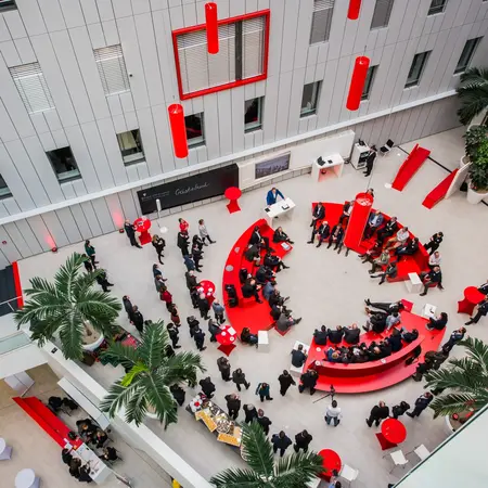 Eine Menge von Menschen versammelt sich in einem modernen, hellen Innenraum mit roten Möbeln und Pflanzen. Die Anordnung der Sitzgelegenheiten bildet eine kreisförmige Struktur, in der verschiedene Gruppen Gespräche führen. Das Ambiente wirkt einladend und kommunikativ.