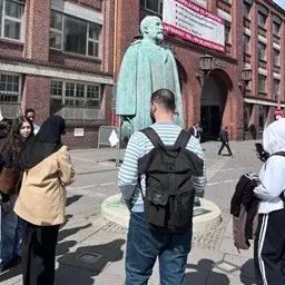 Eine Gruppe von Menschen steht um eine Statue eines historischen Mannes. Die Personen sind unterschiedlich gekleidet und scheinen miteinander zu sprechen oder das Denkmal zu betrachten. Im Hintergrund sind alte Gebäude und ein Banner sichtbar.