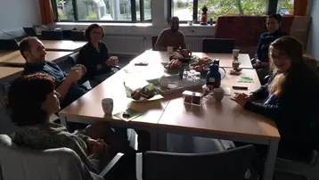 Eine Gruppe von sechs Personen sitzt um einen Tisch, der mit Snacks und Getränken gedeckt ist. Die Atmosphäre ist entspannt und gesellig, während sie miteinander sprechen und ihre Zeit genießen. Heller Raum mit großen Fenstern, durch die Tageslicht strahlt.